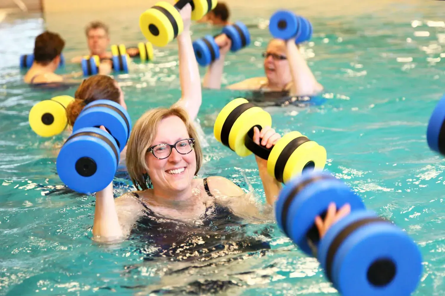 lachende vrouw met bril in het zwembad met blauw/gele gewichten in haar handen